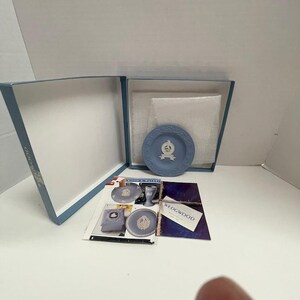 May include: A blue Wedgwood plate with a white emblem inside a blue box. The box is open, revealing the plate, packaging, and printed materials. The Wedgwood logo is visible on the box and printed materials.