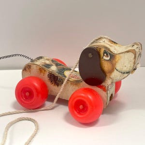 May include: Vintage pull-along toy dog with red wheels. The dog has a brown and white spotted body, a brown ear, and a long, cream-colored pull string. The toy is made of plastic and appears to be well-loved.