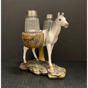 May include: A white horse figurine with a gray mane and tail, carrying two glass jars with silver lids in a brown saddlebag. The horse stands on a rocky base with green accents. A decorative piece.