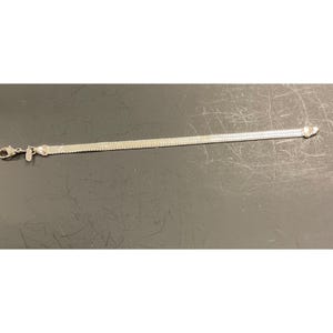 May include: A silver-colored bracelet with a woven chain design. The bracelet has a lobster clasp closure and a small charm near the clasp. The bracelet is lying on a dark surface.