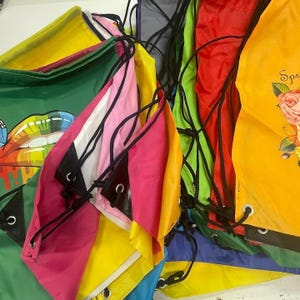 May include: A collection of colorful drawstring bags in various shades, including pink, yellow, green, and red. Some bags feature graphic designs, while others are solid colors. The bags have black drawstrings and eyelets.