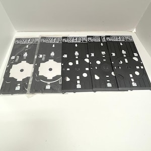 May include: A collection of black plastic trays with various cut-out shapes and white accents. The trays have a rectangular shape with circular and square openings. Some trays are still in a clear plastic wrapping.