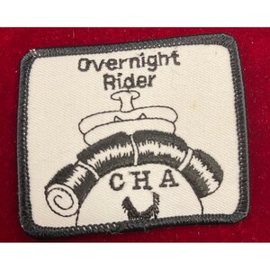 May include: A rectangular patch with a black border and a white background. The patch features the words "Overnight Rider" above a black illustration of a saddle and rolled blankets. The letters "CHA" are below the illustration.