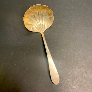 May include: A silver-toned serving spoon with a shell-shaped bowl and a long, slender handle. The bowl has a scalloped edge and a textured, fan-like design. The spoon is likely made of metal and is set against a dark background.
