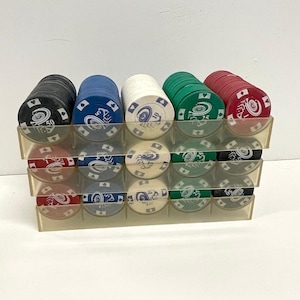 May include: A clear plastic rack holding three rows of stacked poker chips in black, blue, white, green, and red. Each chip features a central roulette wheel graphic, a numerical value, and suit symbols including spades, hearts, diamonds, and clubs.