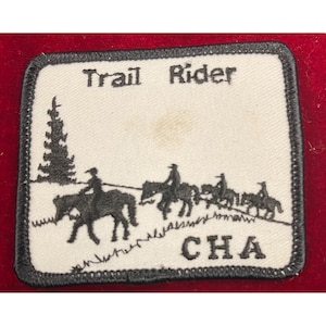 May include: A rectangular patch with a black border and the words "Trail Rider" at the top. The patch features a black silhouette of a pine tree and several riders on horseback, with the letters "CHA" at the bottom.