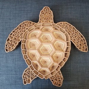 May include: A layered wooden sea turtle sculpture. The artwork features a detailed design with multiple layers of wood, creating a three-dimensional effect. The turtle's shell is composed of hexagonal shapes, and the overall color is a natural wood tone.
