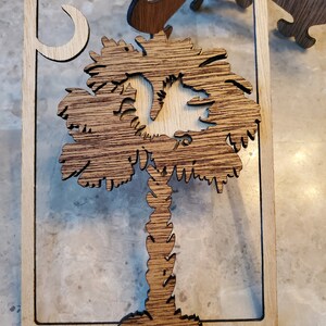 May include: A wooden wall art piece featuring a detailed palm tree design with a crescent moon in the upper left corner. The artwork is made of layered wood, with a light-colored frame and a darker, textured tree.