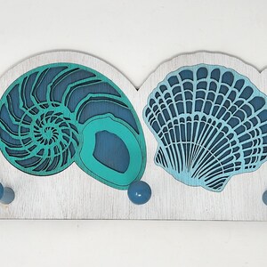 May include: A white wooden coat rack with five hooks. The rack is decorated with five blue and green sea-themed cutouts: a sand dollar, a nautilus shell, a scallop shell, and two starfish.