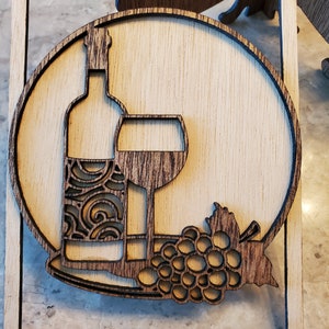 May include: A wooden cutout of a wine bottle, wine glass, and grapes. The design is in a circle and is surrounded by a square frame.