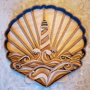 May include: Wooden layered art piece depicting a lighthouse scene. The artwork features a lighthouse with a striped design, waves, and sun rays, all within a shell-shaped frame. The piece is made of various shades of wood.