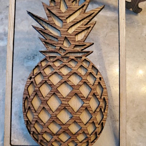 May include: A wooden pineapple cutout with a detailed design. The pineapple is framed in a square wooden frame.
