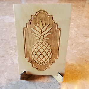 May include: A wooden sign with a carved pineapple design. The pineapple is in a decorative frame and the sign is on a small wooden stand.