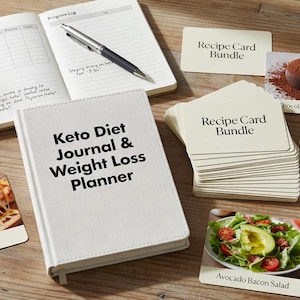 Op de afbeelding: Een witte Keto Diet Journal & Weight Loss Planner ligt op een houten oppervlak, samen met receptkaarten, een notitieboek en een pen. Receptkaarten tonen afbeeldingen van keto-vriendelijke maaltijden zoals pizza en avocado-spek-salade.