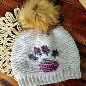 May include: White crocheted beanie with a purple and gray paw print design and a fluffy brown pom pom on top.