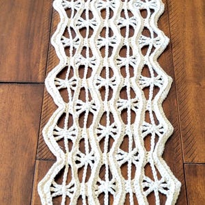 May include: Cream-coloured, handmade crochet table runner with a repeating geometric pattern. The runner has a scalloped edge and is finished with wooden beads and tassels at the ends. It is displayed on a wooden surface.