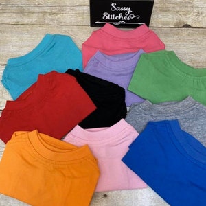 May include: A stack of ten plain t-shirts in various colors, including orange, pink, blue, gray, green, black, red, purple, and turquoise. The shirts are folded and stacked on a wooden surface. The text "Sassy Stitches" is visible on a black sign.