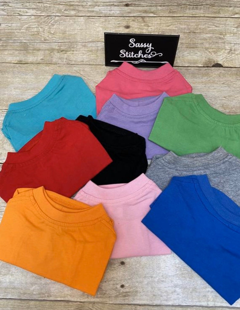 May include: A stack of ten plain t-shirts in various colors, including orange, pink, blue, gray, green, black, red, purple, and turquoise. The shirts are folded and stacked on a wooden surface. The text "Sassy Stitches" is visible on a black sign.
