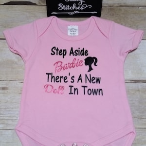 May include: Pink baby onesie with the text "Step Aside Barbie There's A New Doll In Town" in black and pink glitter lettering. A black silhouette of a ponytail is next to the word "Barbie". The onesie has three snaps at the bottom.