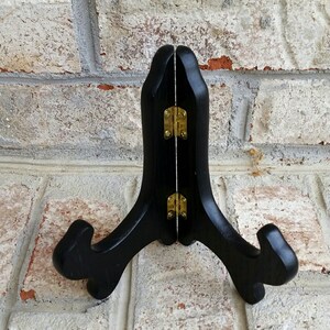 May include: Black wooden easel stand with gold hinges. The stand is designed to hold a plate or other flat object.