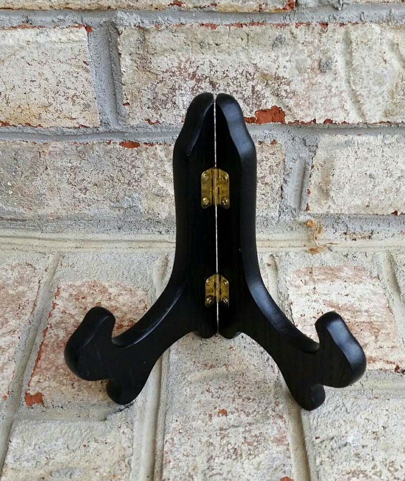 May include: Black wooden easel stand with gold hinges. The stand is designed to hold a plate or other flat object.