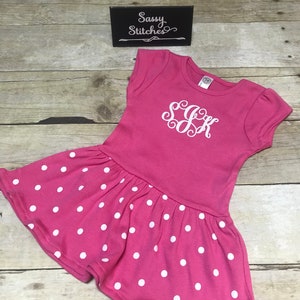 May include: Pink polka dot dress with a white monogram "SGR" on the front. The dress has short sleeves and a ruffled hem. The dress is on a wooden background with a sign that says "Sassy Stitches".