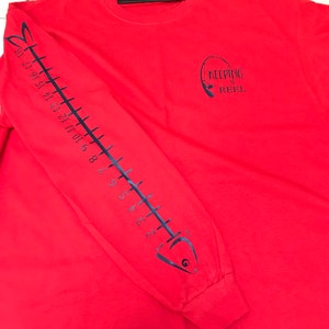 May include: Red long-sleeve t-shirt with a black fish skeleton design on the left sleeve and the text "KEEPING IT REEL" with a fishing rod graphic on the right side of the shirt.