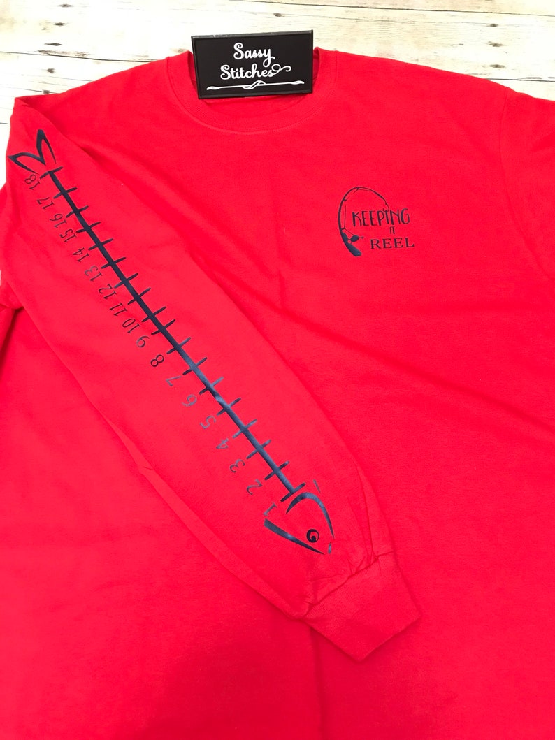May include: Red long-sleeve t-shirt with a black fish skeleton design on the left sleeve and the text "KEEPING IT REEL" with a fishing rod graphic on the right side of the shirt.