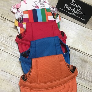 Puede incluir: Cuatro arneses para perros diferentes, apilados uno encima del otro. Los arneses est&aacute;n hechos de diferentes telas y colores, incluyendo floral, a rayas, rojo, azul y naranja. El arn&eacute;s superior es floral, el segundo es a rayas, el tercero es rojo, el cuarto es azul y el arn&eacute;s inferior es naranja. Los arneses son todos para perros peque&ntilde;os. La imagen tambi&eacute;n incluye un letrero que dice "Sassy Stitches".