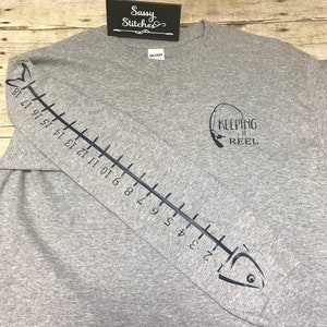 May include: Gray long-sleeve t-shirt with a fish graphic on the sleeve and a fishing rod graphic on the front. The fish graphic has a ruler on its side with numbers 1 through 18. The fishing rod graphic has the text "Keeping it Reel".