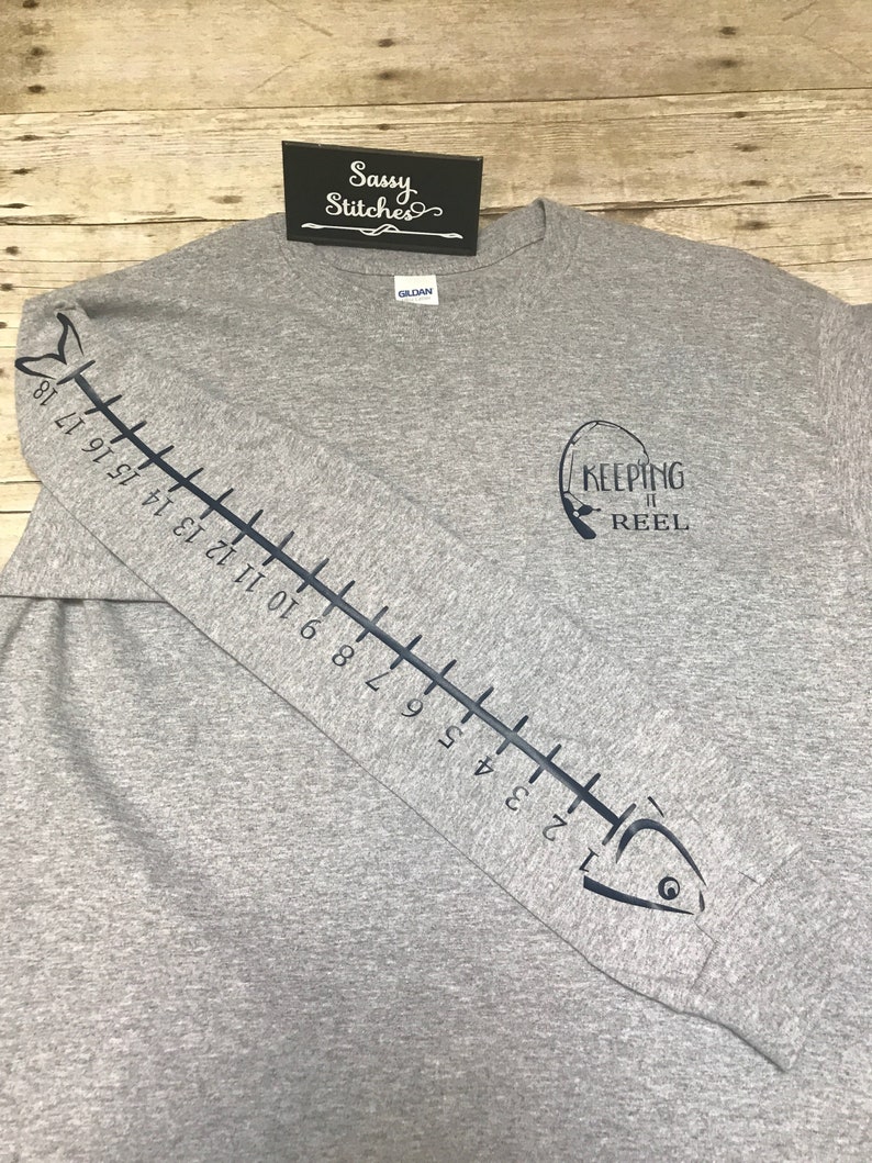 May include: Gray long-sleeve t-shirt with a fish graphic on the sleeve and a fishing rod graphic on the front. The fish graphic has a ruler on its side with numbers 1 through 18. The fishing rod graphic has the text "Keeping it Reel".