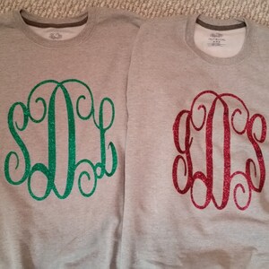 May include: Two gray sweatshirts with a monogram design. One sweatshirt has a green glitter monogram and the other has a red glitter monogram.