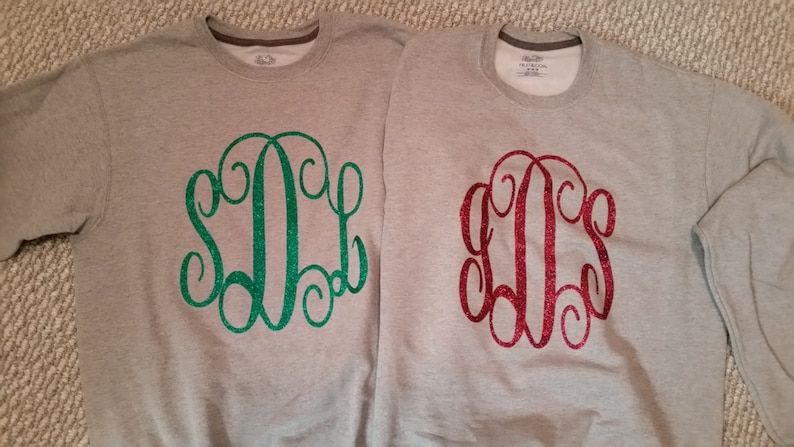 May include: Two gray sweatshirts with a monogram design. One sweatshirt has a green glitter monogram and the other has a red glitter monogram.