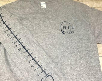 May include: Gray t-shirt with a fish graphic on the sleeve and the text "KEEPING IT REEL" on the front. The fish graphic includes a ruler with numbers 1 through 15.