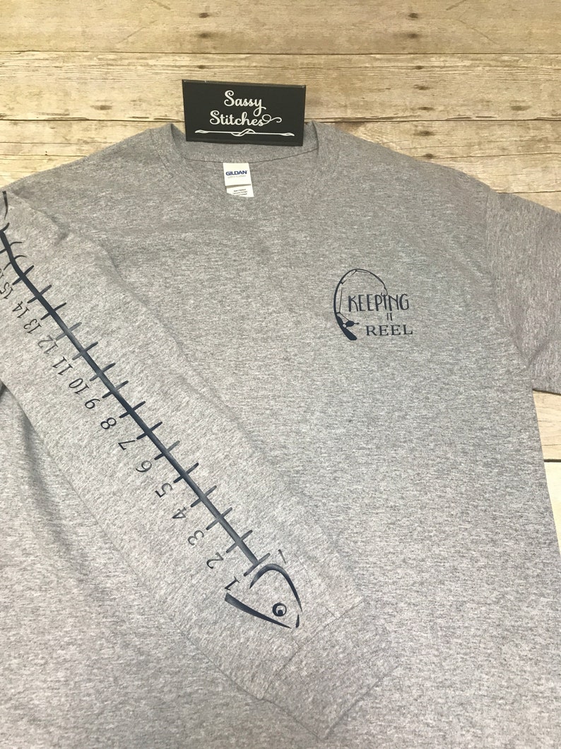 May include: Gray t-shirt with a fish graphic on the sleeve and the text "KEEPING IT REEL" on the front. The fish graphic includes a ruler with numbers 1 through 15.