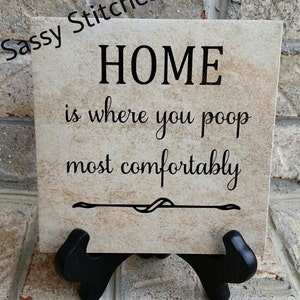 May include: A beige ceramic tile with a black inscription that reads "HOME is where you poop most comfortably". The tile is on a black stand.