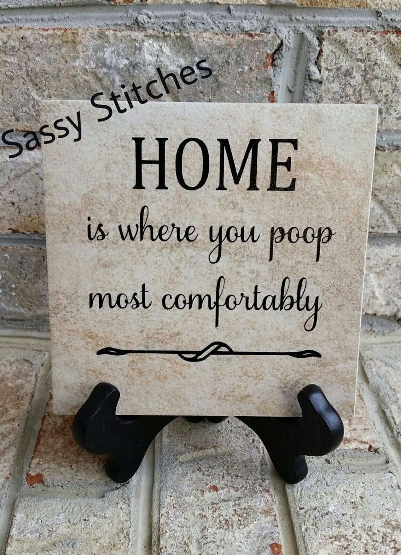 May include: A beige ceramic tile with a black inscription that reads "HOME is where you poop most comfortably". The tile is on a black stand.