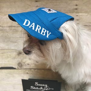 Puede incluir: Un perro blanco con una gorra de b&eacute;isbol azul con el nombre "DARBY" bordado en blanco. La gorra tiene una visera negra y un peque&ntilde;o logotipo en la parte superior. El perro est&aacute; sentado en una superficie de madera con un letrero negro que dice "Sassy Stitches" en blanco.