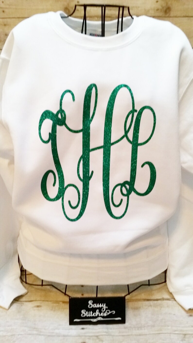 May include: White sweatshirt with a green glitter monogram "SLG" on the front. The sweatshirt is on a hanger and the text "Sassy Stitches" is visible on a black sign below.