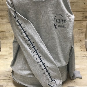 May include: Gray long-sleeve t-shirt with a black fish design on the left sleeve and the text "KEEPING IT REEL" on the right side of the shirt.