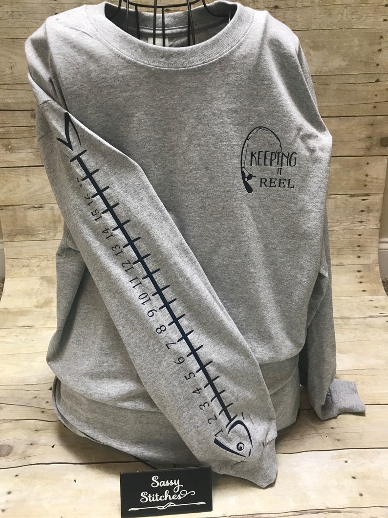 May include: Gray long-sleeve t-shirt with a black fish design on the left sleeve and the text "KEEPING IT REEL" on the right side of the shirt.