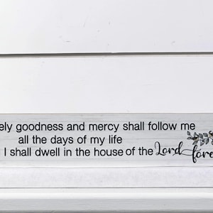 May include: A rectangular wooden sign with the text "Surely goodness and mercy shall follow me all the days of my life and I shall dwell in the house of the Lord forever" and "Psalm 23:6". The sign has a white and gray wood grain pattern.