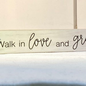 May include: A rectangular wooden block with the words "Walk in love and grace" in black script. A bluebell flower is painted on the left side. The block has a distressed, off-white finish, giving it a rustic appearance.