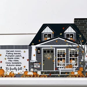 May include: A decorative wooden house with a dark gray exterior, black roof, and a brick chimney. The house is decorated with pumpkins, fall leaves, and the text "It's finally fall". The text also includes "Harvest moon, Falling leaves, Pumpkin patch, Apple cider, Hay rides, Cozy sweaters, Autumn skies."