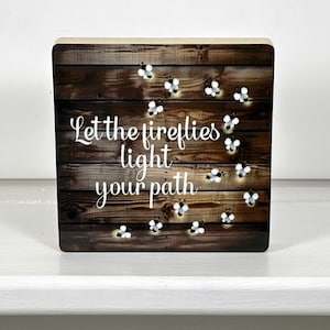 May include: A wooden sign with a rustic brown background and white text that reads "Let the fireflies light your path". The sign is decorated with white fireflies.