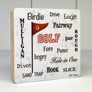 May include: A square wooden block with a white surface featuring golf-related words like "Birdie," "Fairway," and "Hole in One." A red flag with the number 9 and the word "GOLF" are also present. The block has a light brown edge.