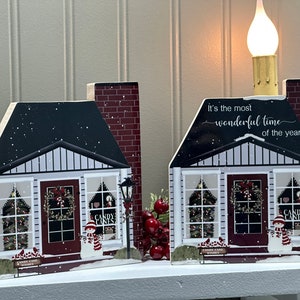 May include: Two wooden Christmas house decorations with red brick chimneys, snow-covered roofs, and windows with Christmas trees inside. The house on the right has the text "It's the most wonderful time of the year" on the roof. Both houses have a snowman in front of them.
