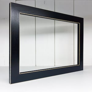 May include: A black picture frame with a thin gold inner border. The frame is rectangular and has a matte finish. The frame is empty, with a white background visible through the opening.