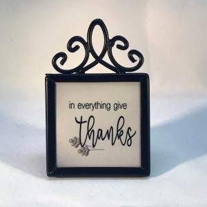 May include: A black metal framed sign with the words "in everything give Thanks" in a script font. The sign has a decorative scroll design at the top and a small floral illustration at the bottom.