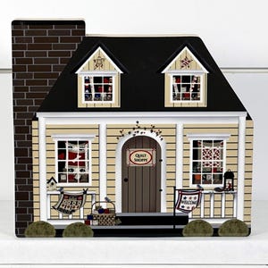 May include: A decorative wooden house with a black roof, tan siding, and a brick chimney. The house features windows with quilt displays, a door with a "Quilt Shoppe" sign, and decorative porch elements. The overall theme is a quaint, country-style shop.